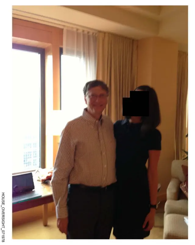Bill Gates stands in a room with a woman. He is smiling, wearing a checkered buttondown. The woman's face is blacked out, but she is wearing black dress