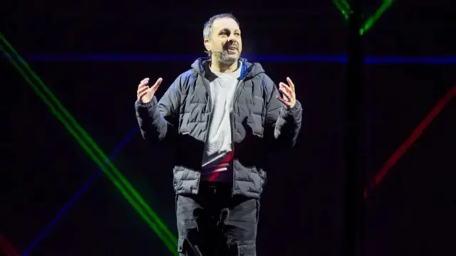 The illusionist Stephen Frayne, wearing a black coat and stood on a stage.