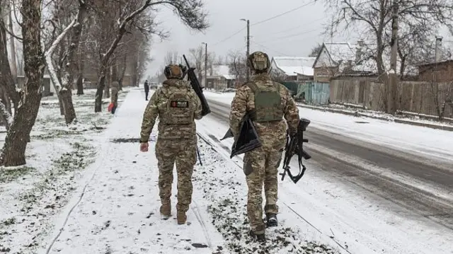 Two Ukranian soldiers holding guns walk on a snow covered street. They are facing away from the camera