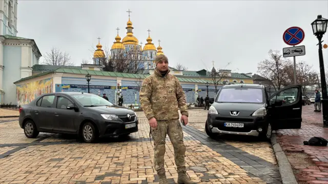 Oleksiy Ostapovych wears camouflage fatigues and stands in front of two cars in a Kyiv street.