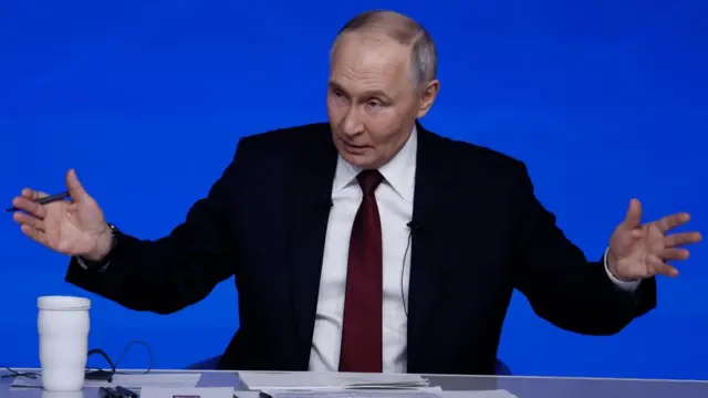 Russian President Vladimir Putin gestures with his arms outstretched as he sits behind a desk and against a blue background.