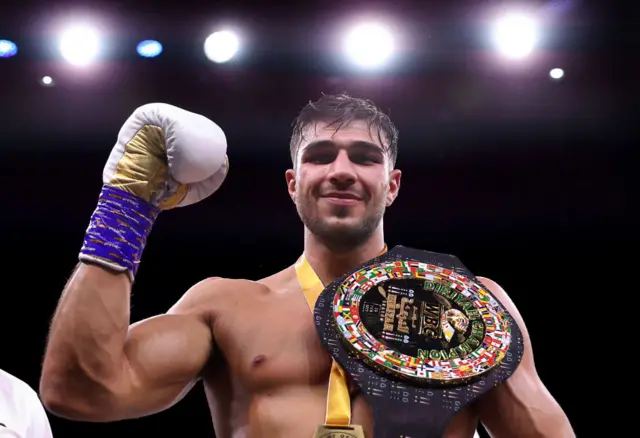 Tommy Fury holds up his boxing glove and has a belt over his shoulder