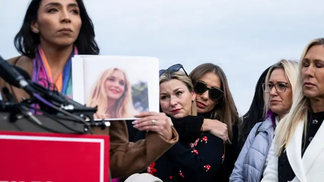 women gathered at a podium, including Epstein survivors, with one holding up a photo of a young survivor