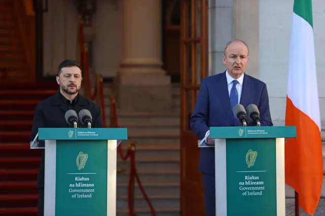 Volodymyr Zelensky in black outfit stands behind a podium bearing the symbol of the Irish Republic on green background, To his left is Irish PM Micheal Martin in a dark blue suit, white shirt and blue tie standing behind a matching podium, an Irish flag behind him