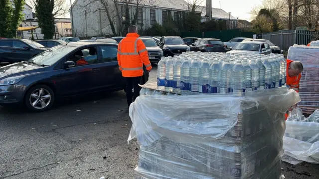 A pallet of bottled water in a car park. A dark-coloured car has pulled up next to it and a man in an orange high-vis jacket is stood behind the pallet.