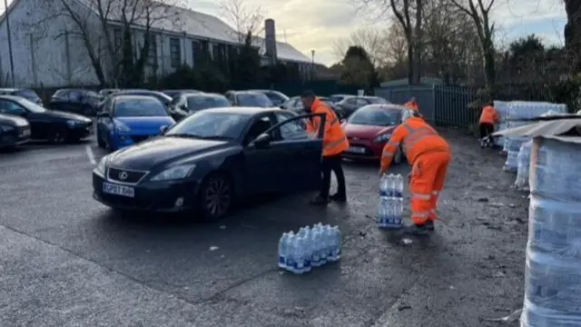 Cars in a car park. Men in high-vis orange clothes are loading bottled water into a car with an open door.