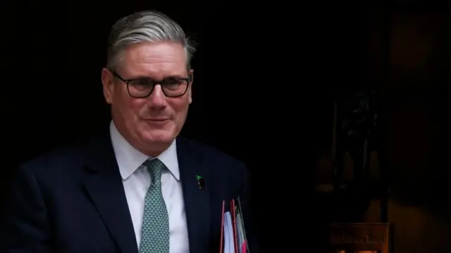 Sir Keir Starmer with a neutral expression emerging from a doorway. he is wearing a suit and carrying two files under his arm.