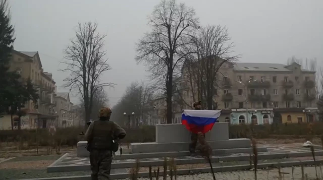 Two soliders wearing camouflage are seen in a town square one of them holding the Russian national flag