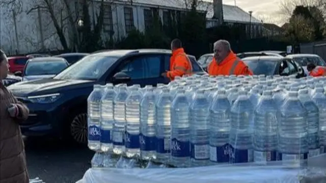 Stacked up cases of bottled water. Behind the stack is  rows of cars and people walking around wearing high-vis clothes