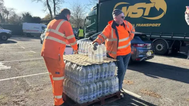Two men in hi vis outfits with crates of water in a car park