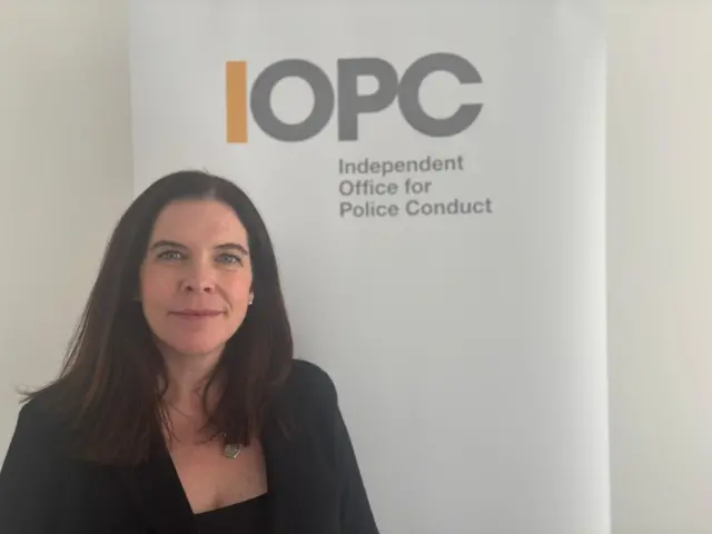 Kathie Cashell, who has long brown hair and wears a black blazer over a black top, smiles at the camera in front of a sign reading IOPC.