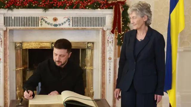 Zelensky writing in a grand book while stood next to Connolly. There's a fireplace with Christmas decorations next to a Ukrainian flag in the background