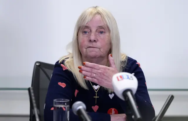 Margaret Aspinall, a middle aged woman with long straight hair and a fringe, gestures with her left hand as she addresses the press. She's wearing a blue jumper with a red hearts pattern and a gold crucifix necklace