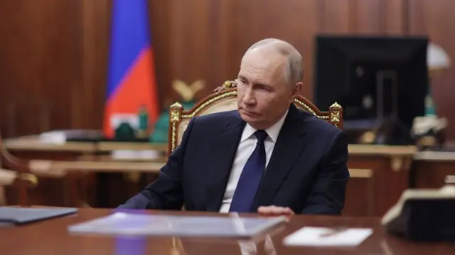 Vladimir Putin in a dark blue suit, matching tie and white shirt sits on a wooden chair with cream brocade cushions. His left hand is lightly gripping a wooden table in front of him, a wooden desk flanked by a Russian flag visible in the background