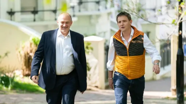 Sir Ed Davey MP and Mike Martin MP walking on a hilly street. Sir Ed is wearing a navy suit and Mike is wearing a yellow and black body warmer.
