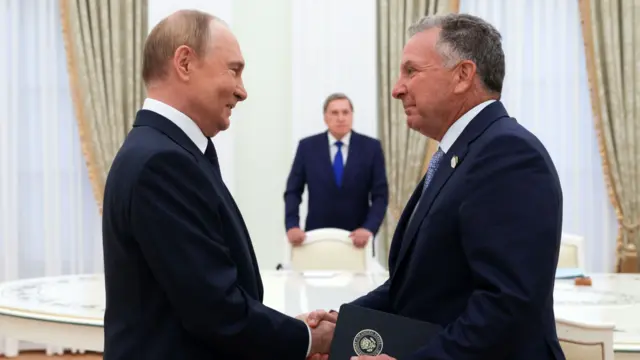 Vladimir Putin (L) smiles as he shakes hands with Steve Witkoff (R). They're both wearing dark suits, there's a white marble-top table surrounded by white chairs behind them. A man in a suit leans on one of the chairs in the blurred background