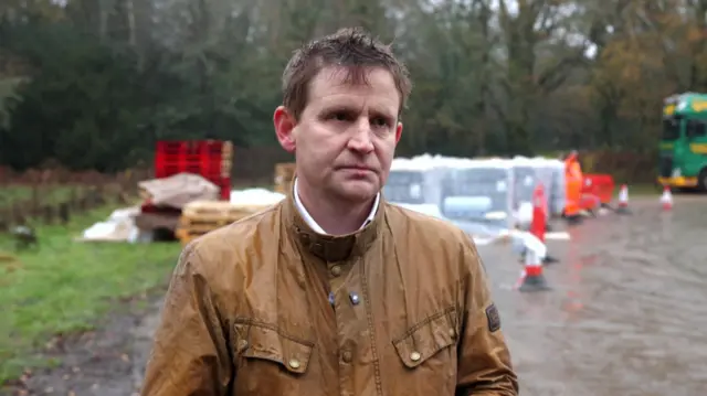 Mike Martin MP wearing a brown jacket, with a stern expression. Pallets of bottled water and some red barriers and traffic cones are seen in the background.