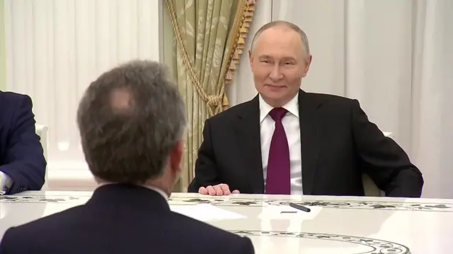 Putin smiling across the table at Witkoff, whose back of head is in the foreground