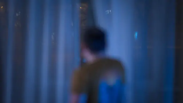 An out of focus image of a man facing away from the camera out of the window through a crack in the curtains.
