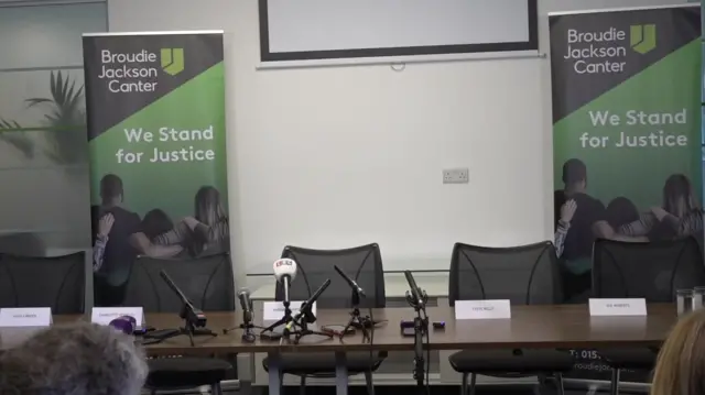 Table with name plates on and microphones pointing towards empty seats. There are two banners behind them reading: 'Broudie Jackson Canter. We Stand for Justice.'