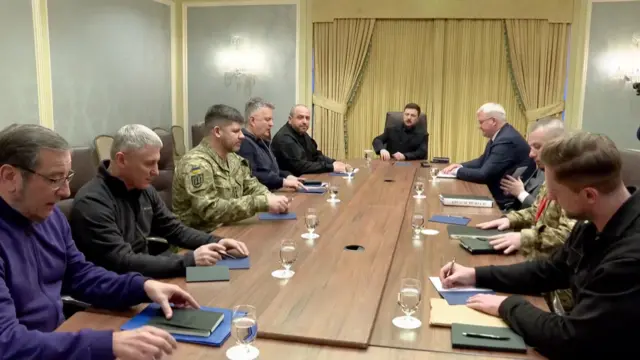 Volodymyr Zelensky, in black clothing, sits at the head of a wooden table surrounded by Ukraine's peace delegation. To his right is delegation head Ruste Umerov, also in black clothing