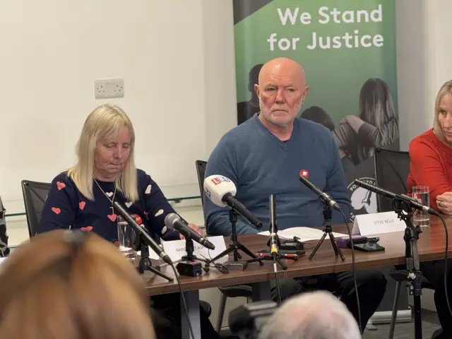 Margaret Aspinall and Steve Kelly sit with several mics in front of them behind a wooden desk