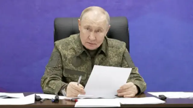 Vladimir Putin in military fatigues sitting at a paper-covered wooden table. He's holding a piece of paper and pen in his hands, a blue wall behind him