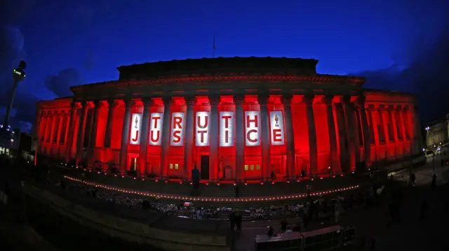A banner reading 'Truth Justice' in red lettering hanging on the pillars of St George's Hall in Liverpool, which is illuminated red