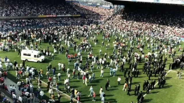 Thousands of fans stream onto the pitch at Hillsborough Stadium. On the right of the frame, by the half-way line,is a line of police officers in black uniform, and on the right a single ambulance drives onto the pitch.