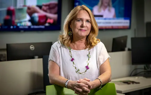 Julie K Brown stands in the Miami Herald newsroom, with her arms resting on a green chair. She is in a white T-shirt and wears a necklace and bracelets.