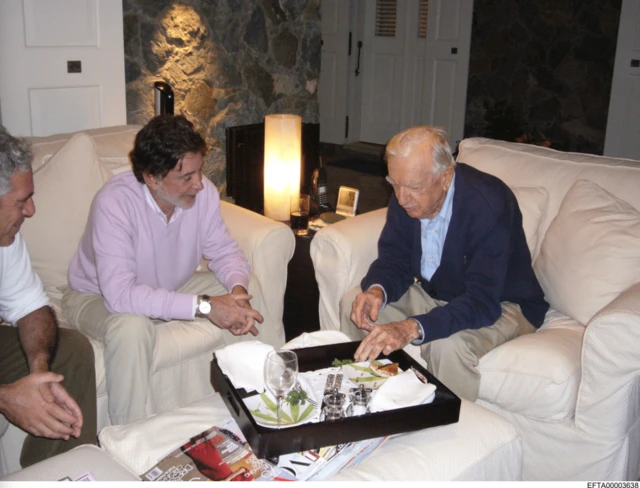Walter Cronkite pictured alongside Epstein and another man, sitting on sofas in living room environment with sandwich tray in front of him