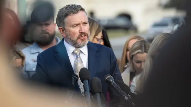 Thomas Massie in a suit speaking into several mics of a lectern outside, he's surrounded by a large group of women