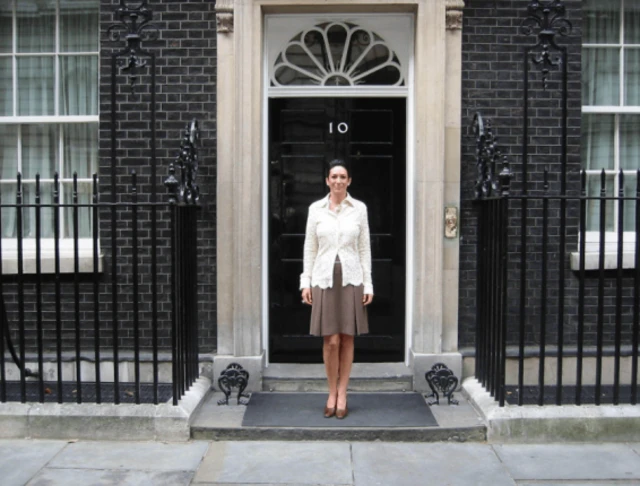 Ghislaine Maxwell stands in front of No 10. She wears a white shirt, brown skirt and brown shoes.