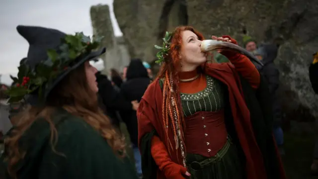 A woman drinks from a horn next to the stones