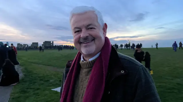 Win Scott wearing a brown umper, dark grey jacket and red scarf. He is standing in a field at sunrise, smiling at the camera with Stonehenge visible behind him.