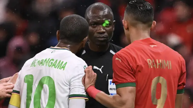 Referee Jean-Jacques Ndala has a green laser shone into his eyes as he is surrounded by a Comoros and a Morocco player during the opening match of Afcon 2025
