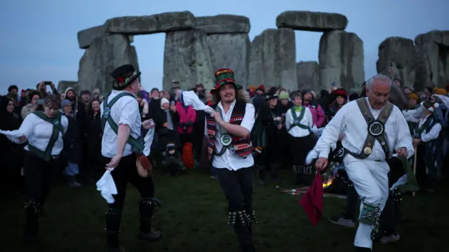 Morris dancers perform as people attend winter solstice celebrations at Stonehenge stone circle. The sun is gradually rising but the sky is still a cloudy blue.