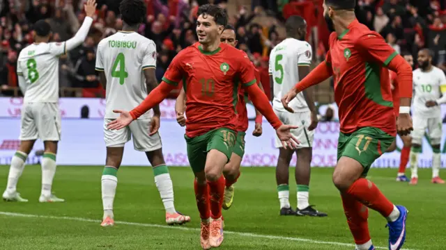 Brahim Diaz in red and green Morocco kit spreads his arms out either side as he runs in celebration, being chased by two team-mates in the opening match of Afcon 2025. Several Comoros defenders in all-white are seen standing in the background, one with his arm up in an appeal