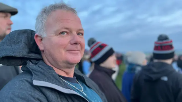 Bradley O'Neill wearing a dark grey hooded jacket and teal t-shirt. He has light grey hair and blue eyes, and is looking at the camera with a slight smile.