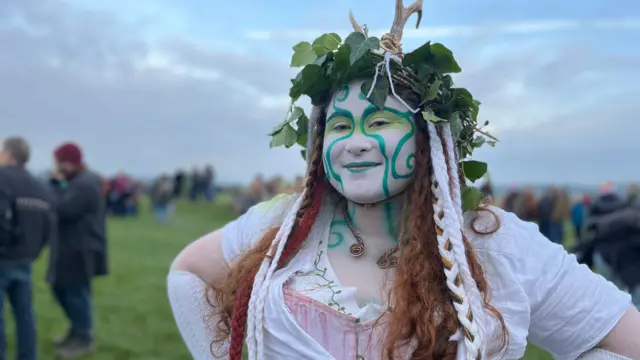 Sophie McCarthy wearing a pink corset, white blouse and a large headdress with white ropes, green leaves and antlers. Her face has been painted white with green eyeshadow and organic wiggly lines.