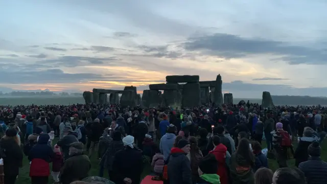 A large crowd of people gathered around the base of Stonehenge. The sun is starting to rise, and the cloudy horizon is turning slightly orange.