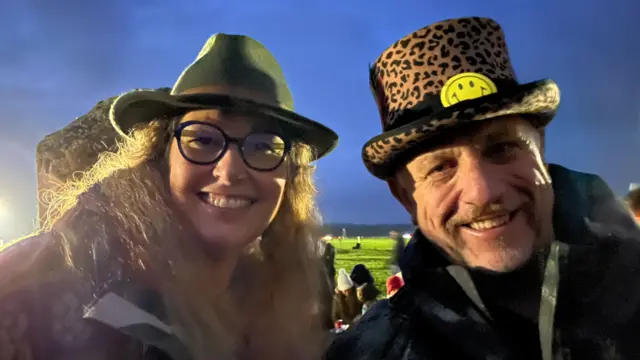 A selfie of Kim Tester and Paul Ransom wearing rain coats and brimmed hats, smiling at the camera. The image is taken just before sunrise when the sky is still a dark blue.
