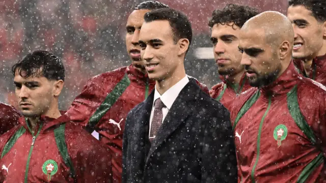 Morocco's Crown Prince Moulay Hassan stands in a big dark coat with shirt and tie, surrounded by members of the Morocco football team in red tracksuit tops with visible rain drops falling
