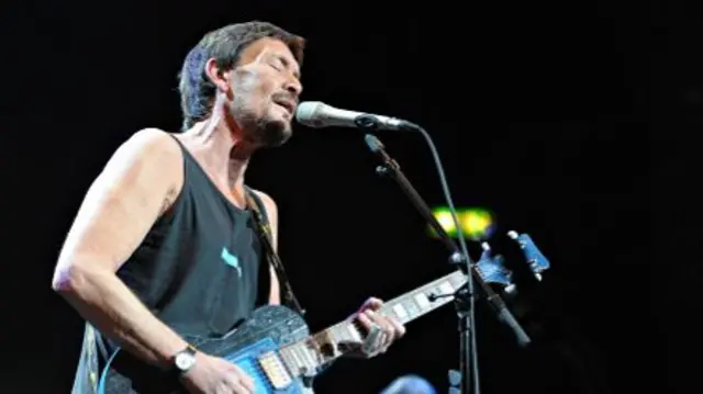 Chris Rea sings into a microphone with his eyes closed as he plays a black electric guitar. He is wearing a black vest