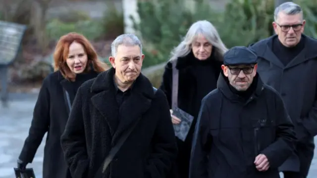 Two men in black jackets stand in the foreground, a row of three people behind them