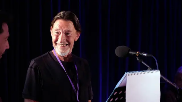 Chris Rea stood in front of a music stand with a microphone in front of him. He has his hair tied back and is smiling