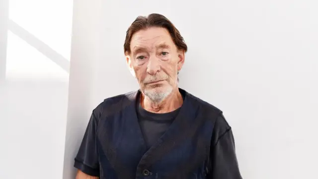 Chris Rea, pictured wearing a blue and black waist coast, standing in front of a white background