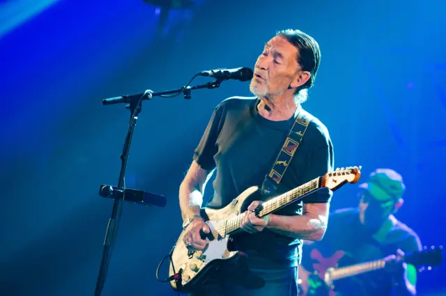 Chris Rea performing with a guitar on stage - there is a microphone in front of him and another guitarist behind him, they are lit in blue light
