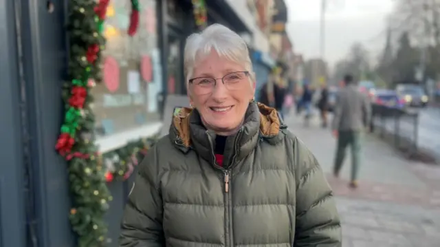 A woman with short grey hair in a green puffer jacket stands on a street