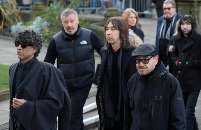 Bobby Gillespie stands in the centre of a group of people in a black coat